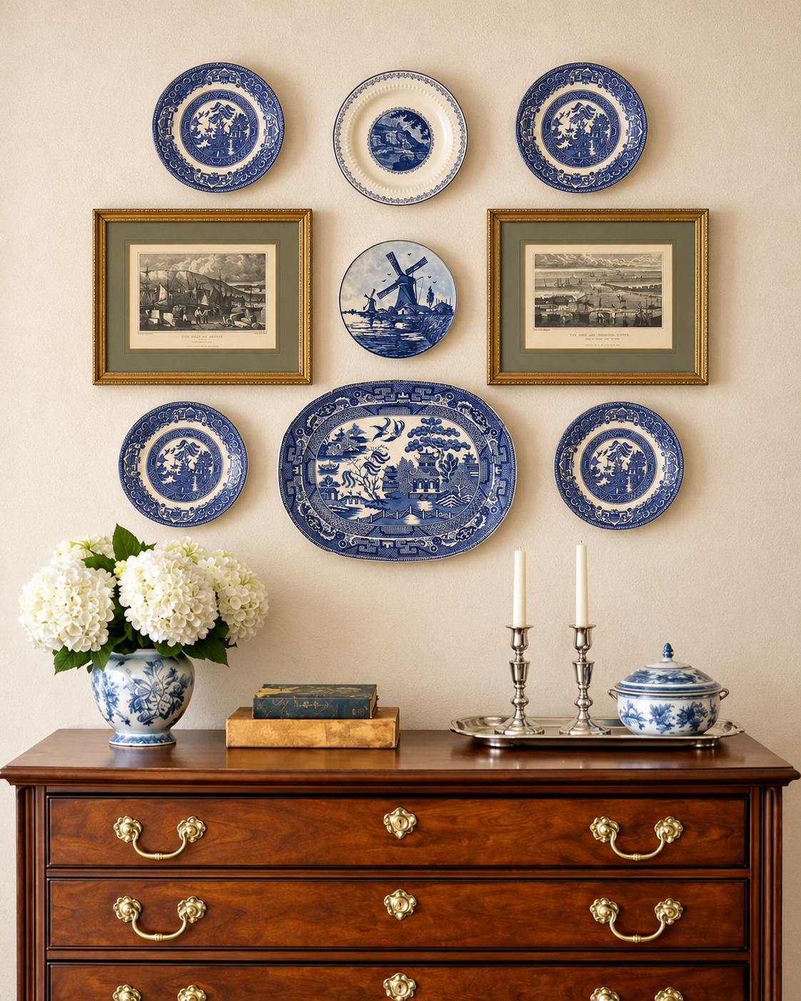 A styled interior scene showing a cream wall decorated with multiple blue-and-white ceramic plates and two framed landscape prints, above a dark wood dresser with brass handles. A blue-and-white floral vase with white flowers, books, candlesticks, and a blue-and-white lidded jar sit on the dresser.